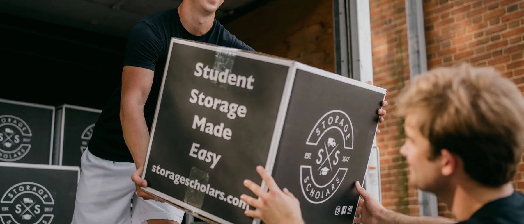 A Storage Scholars crew member hands a branded moving box out of a truck to a student